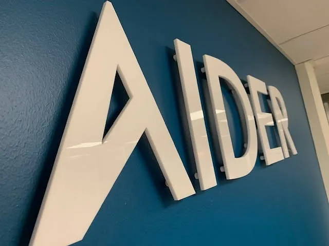 Nærbilde av opphøyd AIDER-logo med hvit finish montert på blå kontorvegg.
