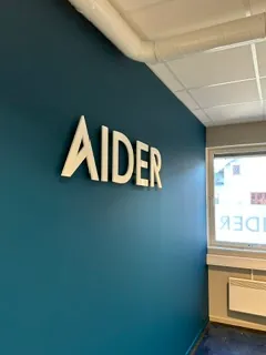 Opphøyde AIDER-bokstaver montert på blå vegg i moderne kontormiljø.