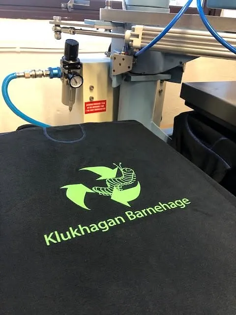 Svart tekstil med logo for Klukhagan Barnehage under produksjon i tekstilpresse.
