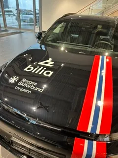 Sort bil med Bilia-profilering, norske fargestriper og kampanjebudskap på panser.