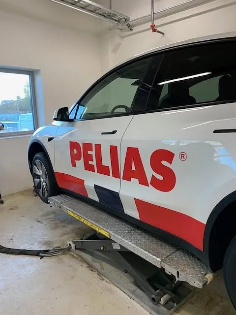 Sidevisning av Tesla med Pelias-profilering på løftebukk under monteringsarbeid.
