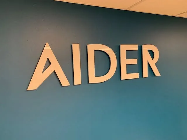 Frittstående AIDER-logo i hvite bokstaver montert på blå vegg innendørs.