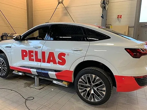 Hvit elbil med Pelias-profilering og dekorstriper, fotografert under montering.
